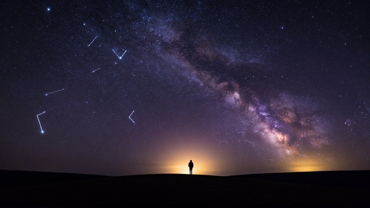 Persona contemplando un cielo nocturno estrellado con un arco de constelaciones sobre una colina oscura.