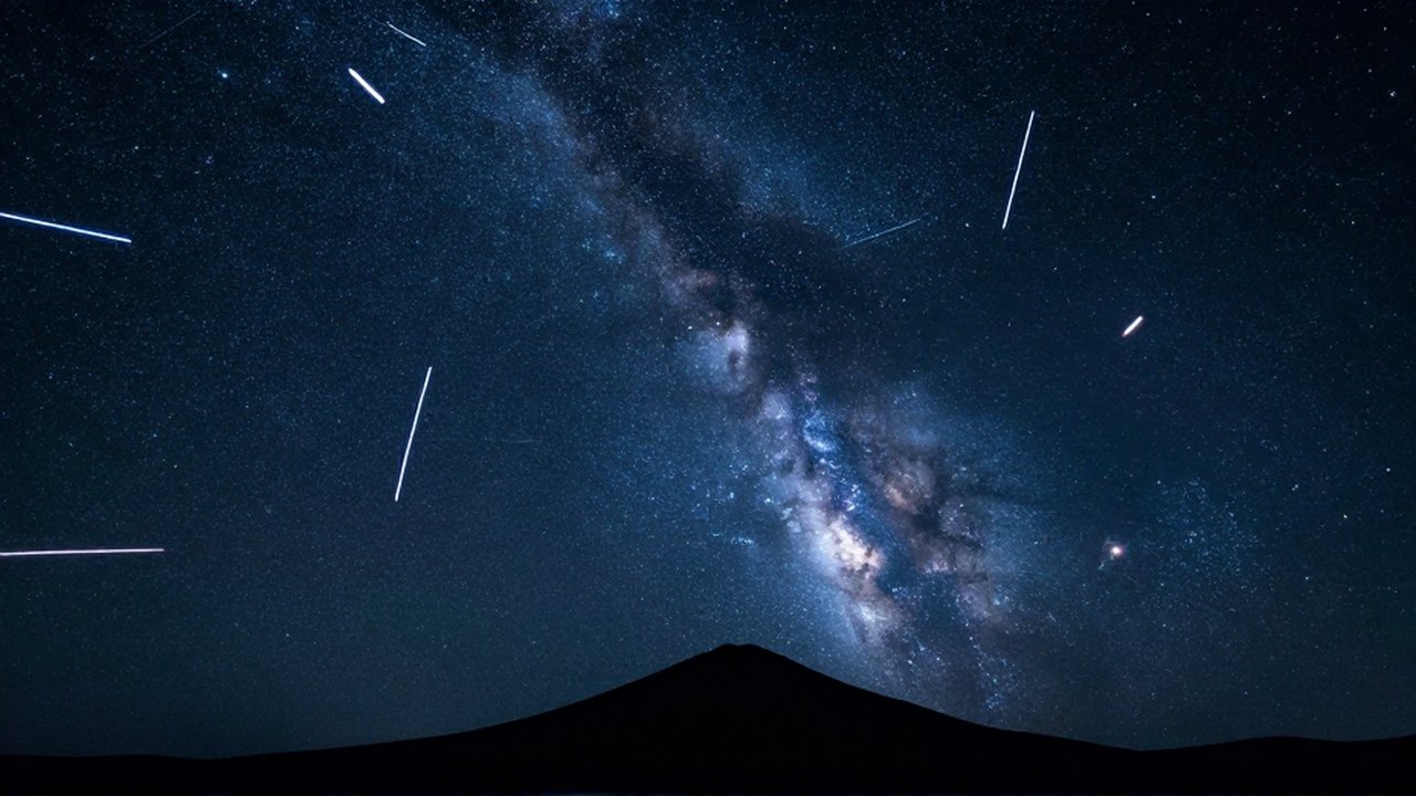 Lluvia de meteoros iluminando un cielo estrellado con la Vía Láctea sobre la silueta de una montaña.